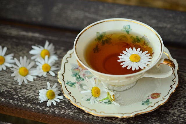 White cup &saucer with light brown tea in it and a floating chamomile blossom and scattered chamomile blossoms on the table.