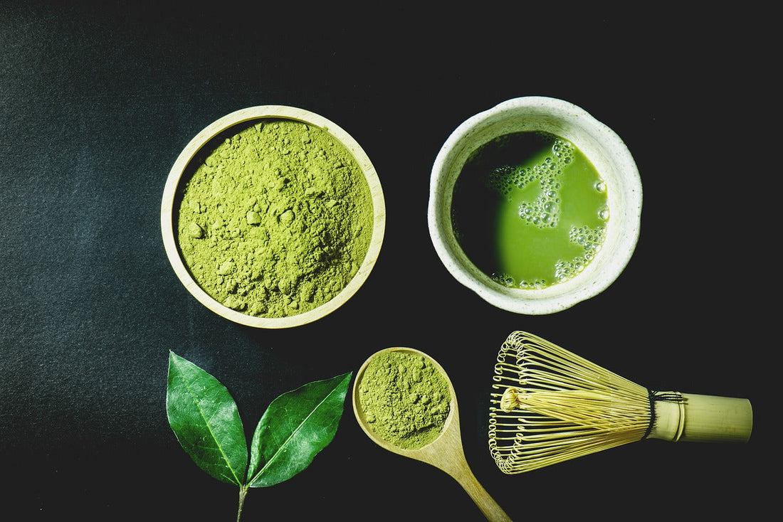 white bowl of dry matcha beside a white bowl of brewed matcha above tea leaves & matcha whisk