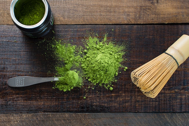 A display of a can of matcha powder and in front matcha powder is scattered on a wood surface with a measuring spoon and a matcha whisk.