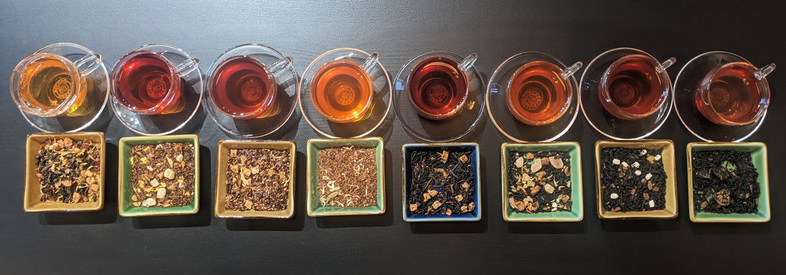8 cups of different colored teas setting on clear saucers, which are setting on a black table with dishes of dry tea leaves in front of them.