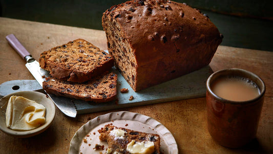 A loaf of sliced Bara Brith setting on a cutting board and surrounded by a knife, butter and a cup of tea