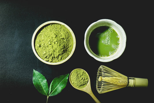 white bowl of dry matcha beside a white bowl of brewed matcha above tea leaves & matcha whisk