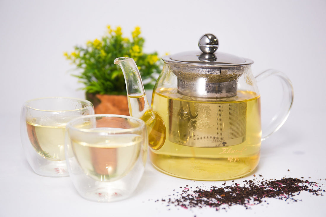 Clear pot with light yellow tea in it and a silver lid/infuser along with two clear cups with very light liquid and a green plant with yellow flowers in a clay pot behind it.